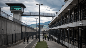 Prison yard with concrete walls, barbed fencing, guard towers, and officers patrolling the walkway.